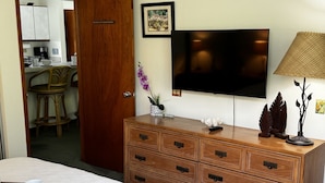 A hotel room with a bed, wooden dresser, TV, and a dining area visible through an open door.