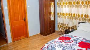 A bedroom with a wooden door, a wardrobe, a bed with a patterned bedspread, and curtains with a geometric design. - Accra Cosy Homes (Ngleshi Amanfro)