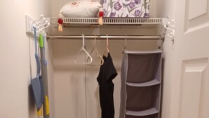 Una camera compatta con un letto, un comodino, un termosifone, una mensola e un appendiabiti con vestiti e prodotti per la pulizia.