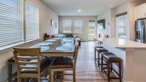 A spacious dining area with a long table, multiple chairs, and a kitchen area with a refrigerator and a TV.