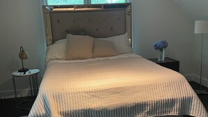 A bedroom with a large bed, a ceiling fan, a small table with a lamp, and a window with a view of trees. - Cozy en-suite in beautiful East Hampton with AC, WiFi (East Hampton)