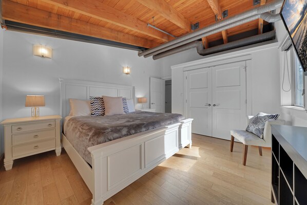 A bedroom with a large bed, white wardrobe, and a desk with a chair. There are two wall-mounted lamps and a ceiling vent.