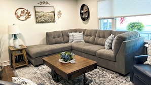 A living room with a sectional sofa, a coffee table with a floral centerpiece, a lamp, and decorative wall art. - Delightful, newly renovated, 3 bedroom cape in a charming country setting  (Marilla)
