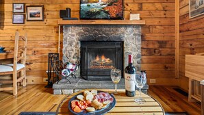 L’intérieur d’un chalet accueillant, avec une cheminée en pierre, des murs en bois, un téléviseur fixé au-dessus de la cheminée, et une table sur laquelle sont disposés des en-cas et une bouteille de vin.