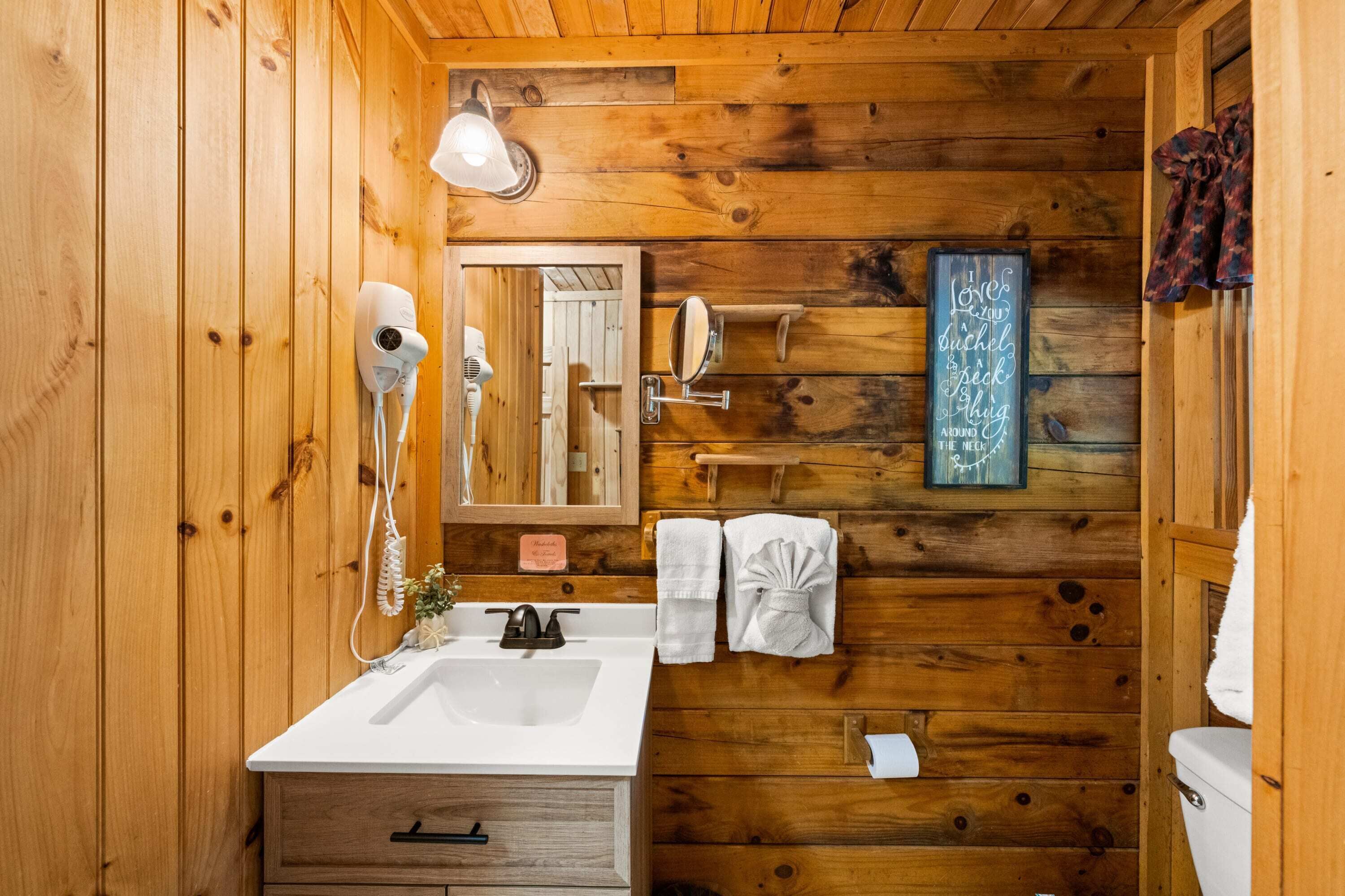 Baño de cabaña de madera con lavamanos blanco, secador de pelo, espejo y toallero.