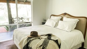 A bedroom with a large bed, white bedding, a hat on the bed, and a view of greenery outside. - Kurraba - Family friendly country escape with pool (Kangaroo Valley)