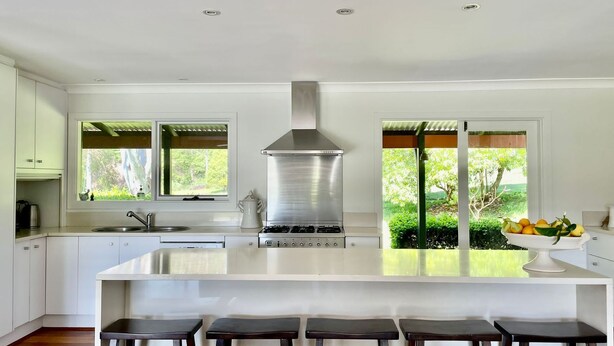 A modern kitchen with a central island, stainless steel range hood, and a view of greenery through large windows. - Kurraba - Family friendly country escape with pool (Kangaroo Valley)