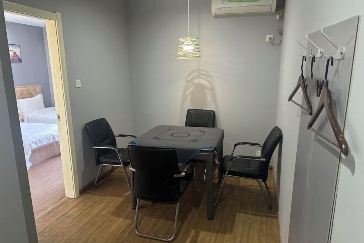 Una habitación pequeña y bien iluminada con una mesa de comedor y cuatro sillas, una cama en la habitación contigua y un aire acondicionado instalado en la pared.