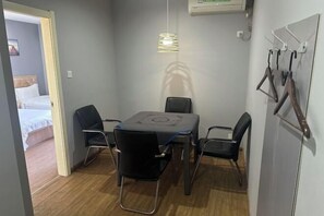 Una habitación pequeña y bien iluminada con una mesa de comedor y cuatro sillas, una cama en la habitación contigua y un aire acondicionado instalado en la pared.