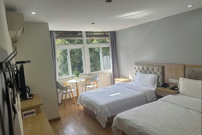 A hotel room with two beds, a desk, a chair, and a window with a view of greenery.