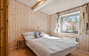 A bedroom with wooden walls, a bed with white bedding, a small stool, and a window with curtains.