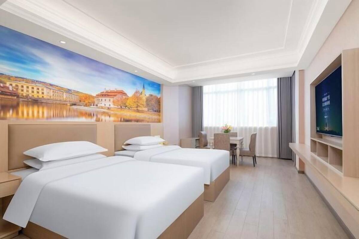 A hotel room with two beds, a desk, a chair, a TV, and a large window.