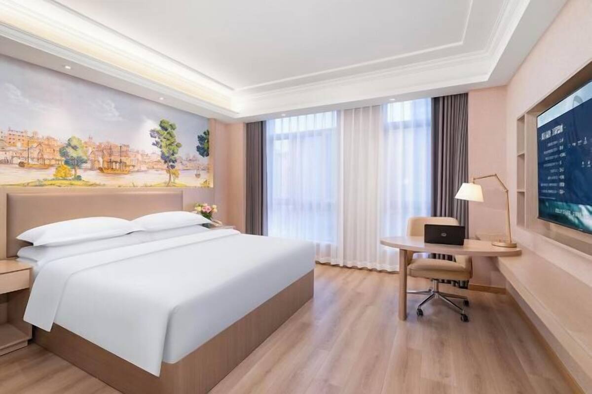 A hotel room with a large bed, a desk with a laptop, a TV, and a mural on the wall.