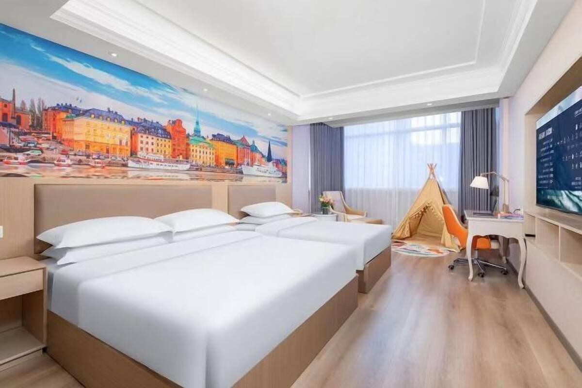 A hotel room with a large bed, a desk, a chair, a TV, and a painting of a cityscape on the wall.