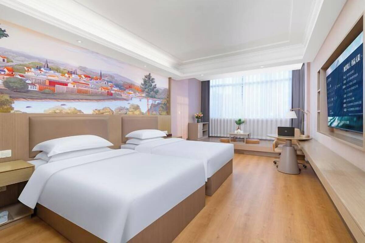 A hotel room with two beds, a desk, a chair, a TV, and a mural on the wall.