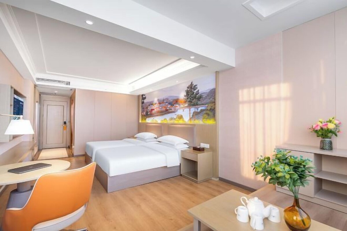 A hotel room with a bed, a desk, a chair, a nightstand, a vase with flowers, and a painting on the wall.