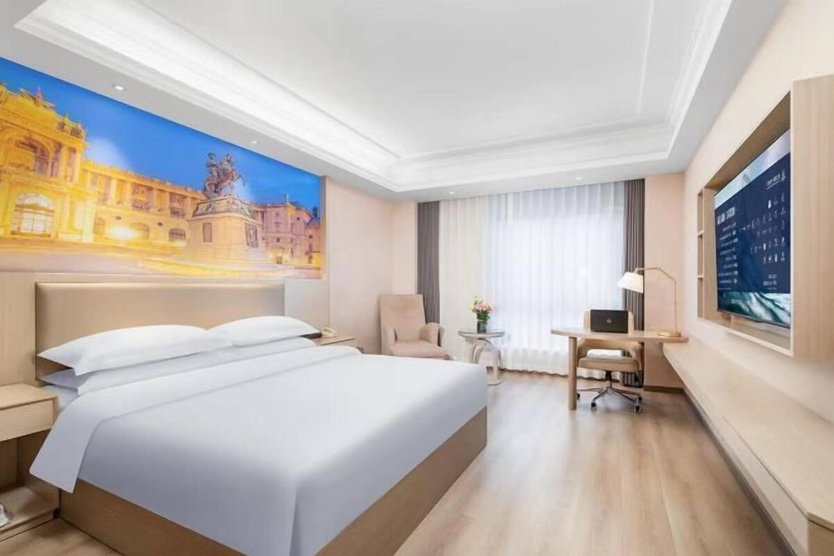 A hotel room with a large bed, a desk, a chair, a small table with a lamp, and a large window with curtains.