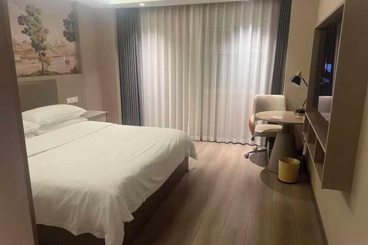 A hotel room with a bed, a desk, a chair, a small table, and a window with curtains.