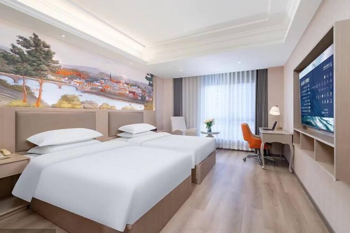 A hotel room with two beds, a desk, a chair, a TV, and a mural on the wall.