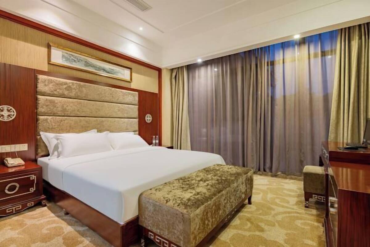 Habitación de hotel con una cama grande, cabecera de madera, mesita de noche con teléfono, un banco y una ventana amplia con cortinas.