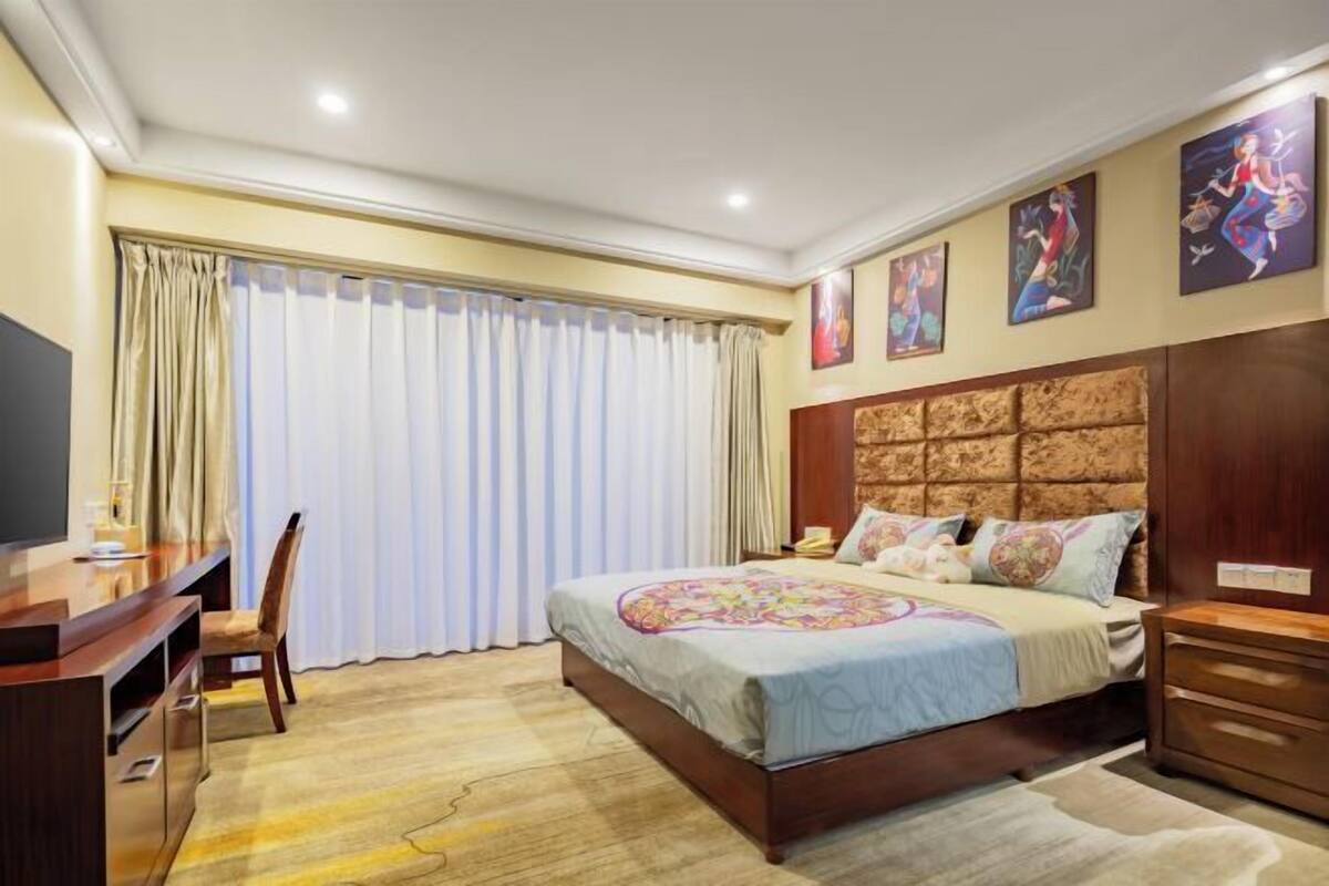 Un dormitorio con cama, escritorio, silla, televisor y obras de arte en la pared.
