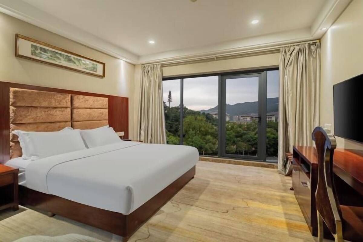 Habitación de hotel con una cama grande, televisión, escritorio y vistas a la ciudad a través de un ventanal amplio.