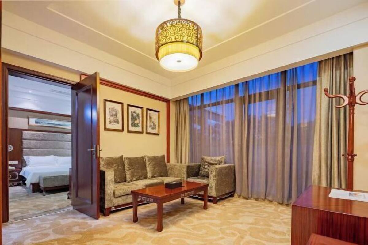 Habitación de hotel con sofá, una mesa de centro, un escritorio y un ventanal con cortinas.