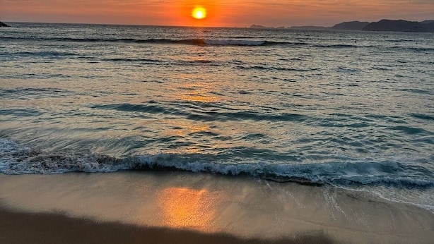 Une plage au coucher du soleil, avec le soleil près de l’horizon, une mer calme et des montagnes lointaines.