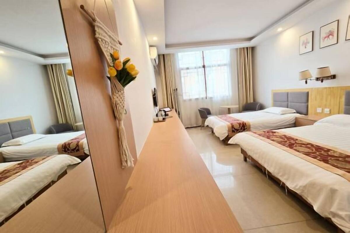 Habitación de hotel con tres camas, una mesa de madera alargada, un espejo y un jarrón con flores.