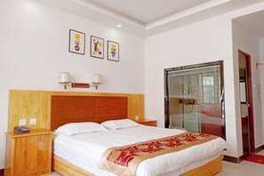 Een hotelkamer met een houten hoofdeinde, een bed met witte lakens, een rode decoratieve plaid, een nachtkastje met een lamp en een glazen deur naar een badkamer.