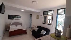 A bedroom with a bed, a black sofa, a small table, and a window with blinds. - Residence KAYA, Marie-Galante, sea view (Grand-Bourg)