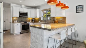 Una cocina moderna con armarios blancos, encimeras de granito y electrodomésticos de acero inoxidable, además de una isla central con taburetes de bar.