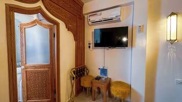 A room with a wooden door, an air conditioning unit, a flat-screen TV, a small wooden table, and a stool.
