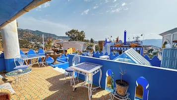 Een terras in blauwtinten, buiten, met een tafel, stoelen, plantenbakken en een kasteelachtige constructie op de achtergrond.