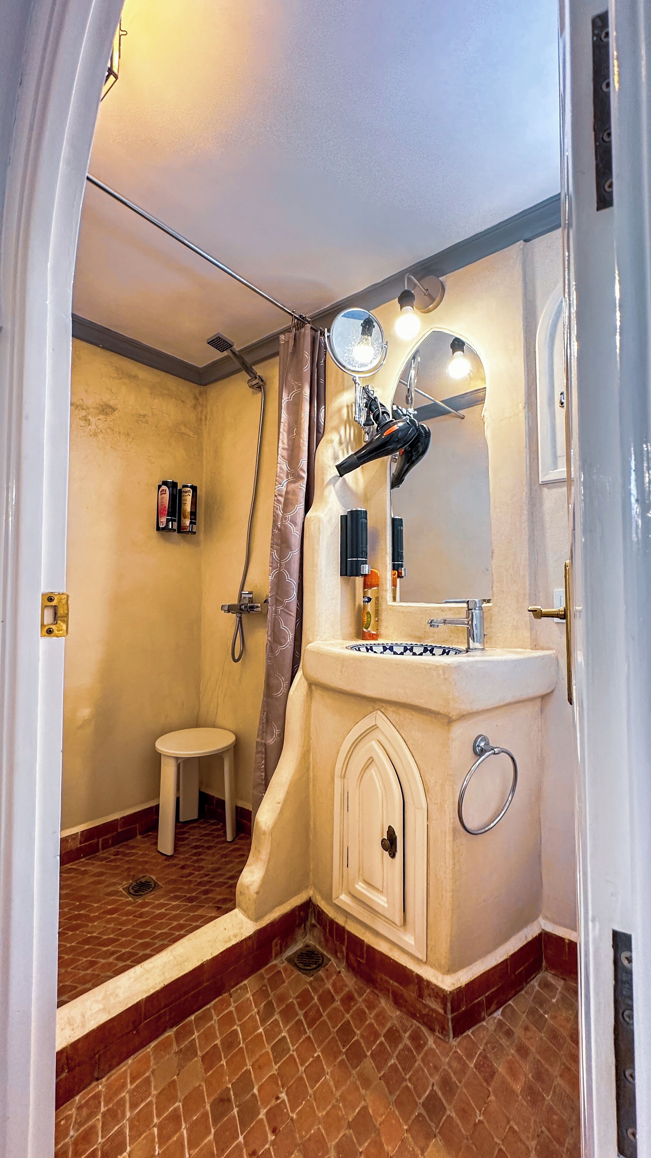 Ein kleines Badezimmer mit Dusche, einer kleinen Bank, einem Waschtisch mit Spiegel und einem Handtuchhalter.