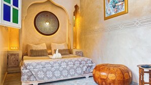Een slaapkamer met een bed, kussens, een decoratieve wand, een houten poef en een klein tafeltje.