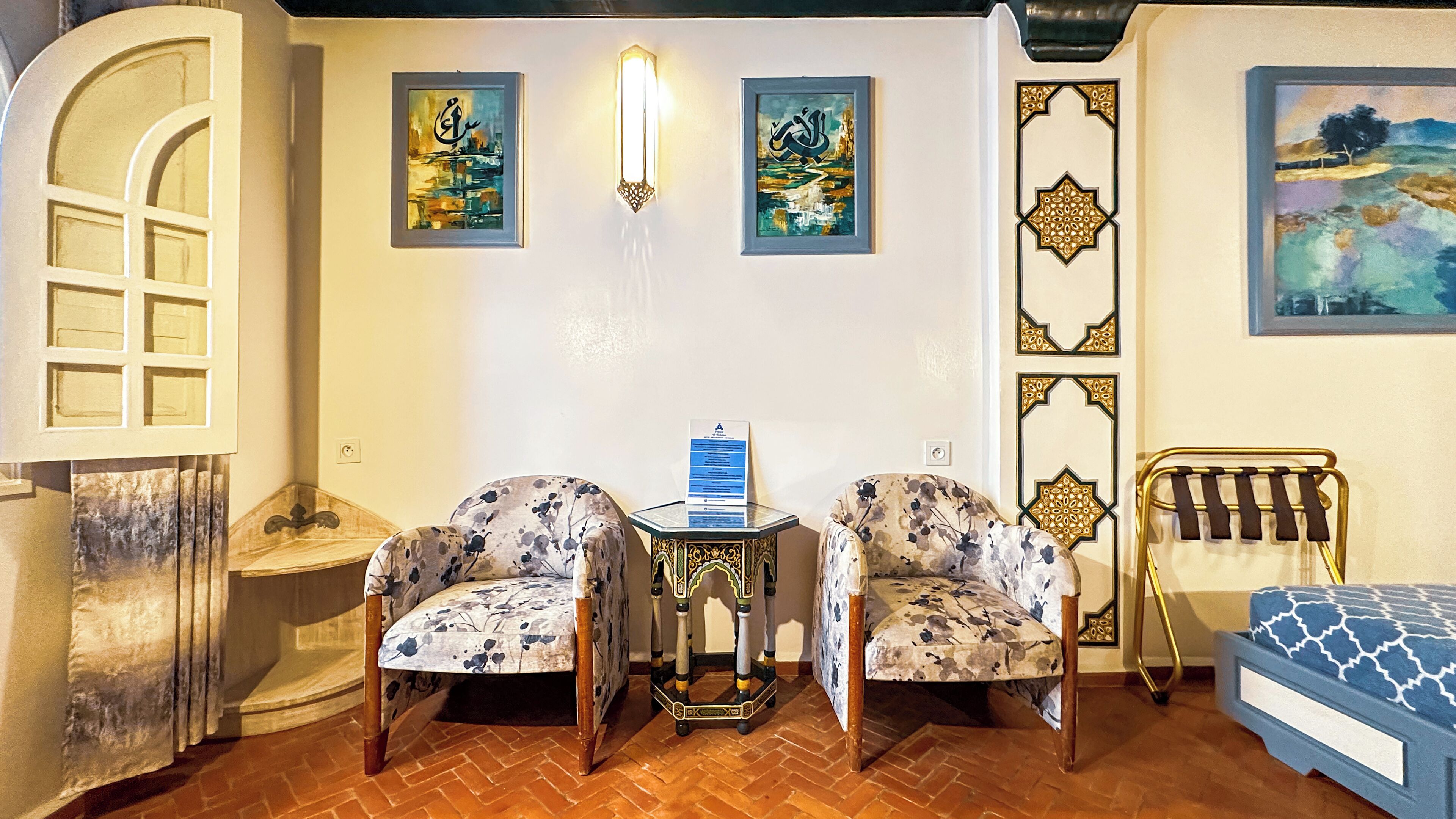 Una habitación con dos sillas de estampado floral, una mesita con un letrero, una cama con colcha estampada y arte decorativo en la pared que incluye caligrafía árabe y diseños geométricos.