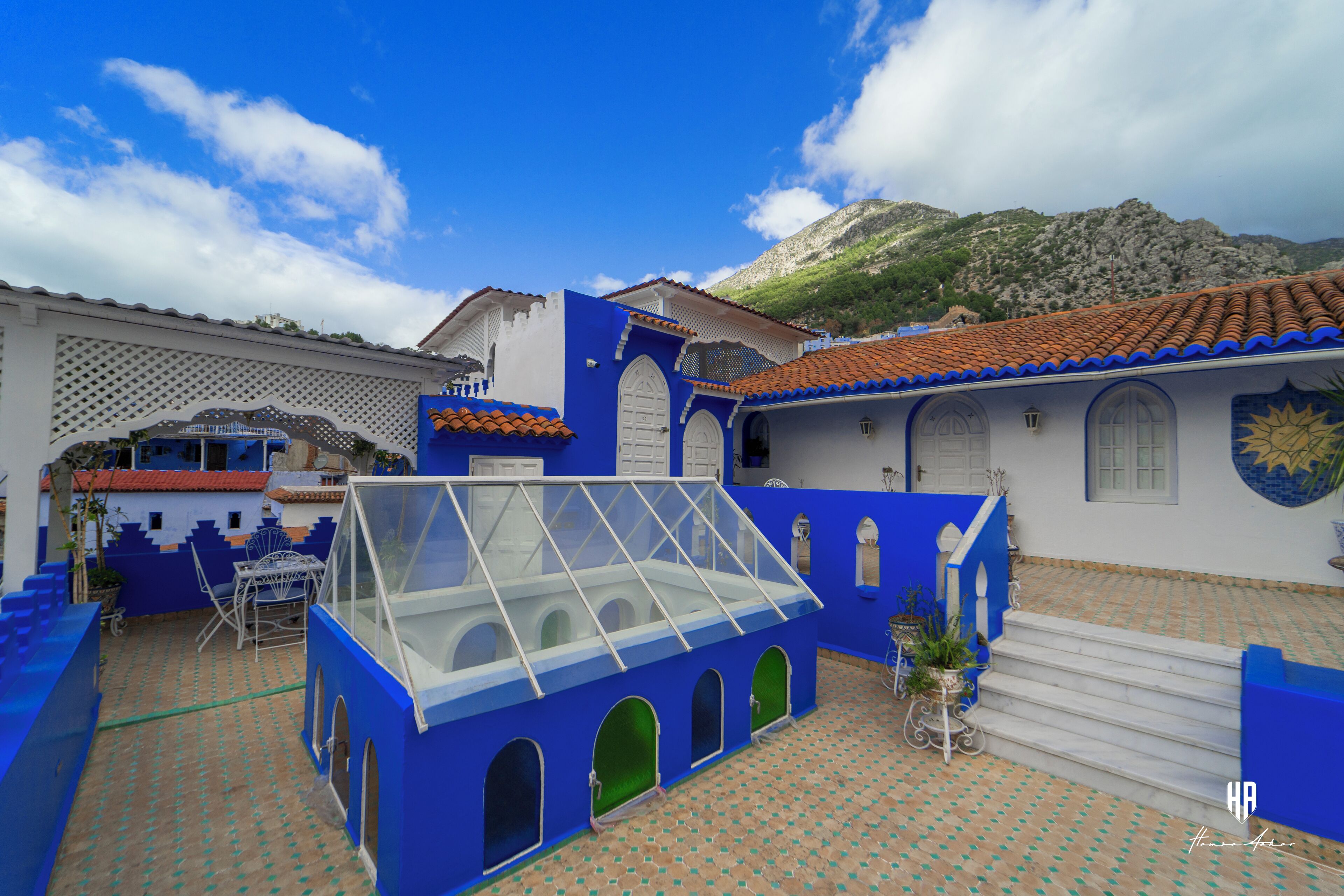 A courtyard with blue and white buildings, a glass-roofed structure, and a mountain in the background.