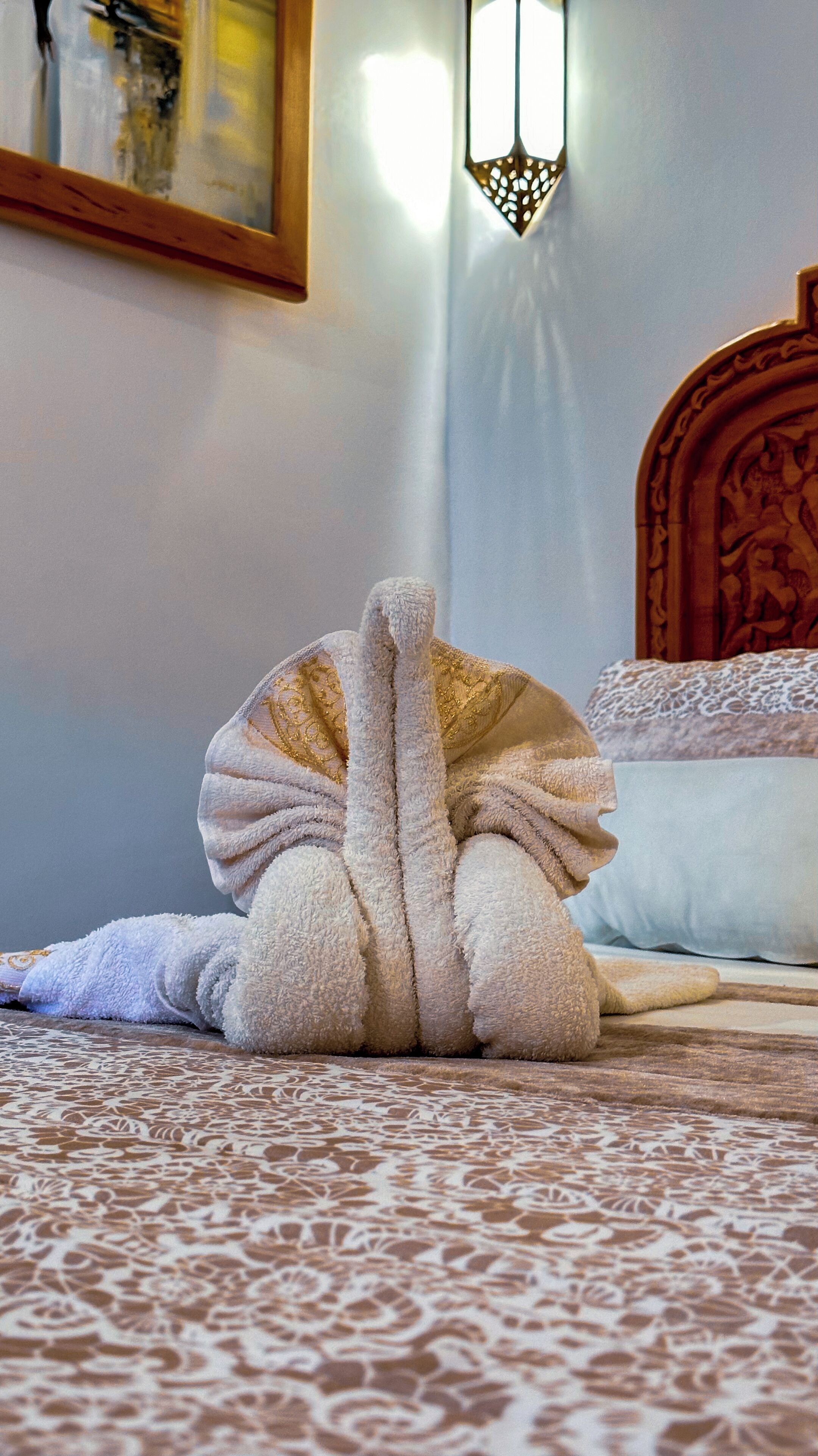 Una toalla doblada cuidadosamente sobre una colcha estampada en una habitación con cabecera de madera y una lámpara fijada a la pared.
