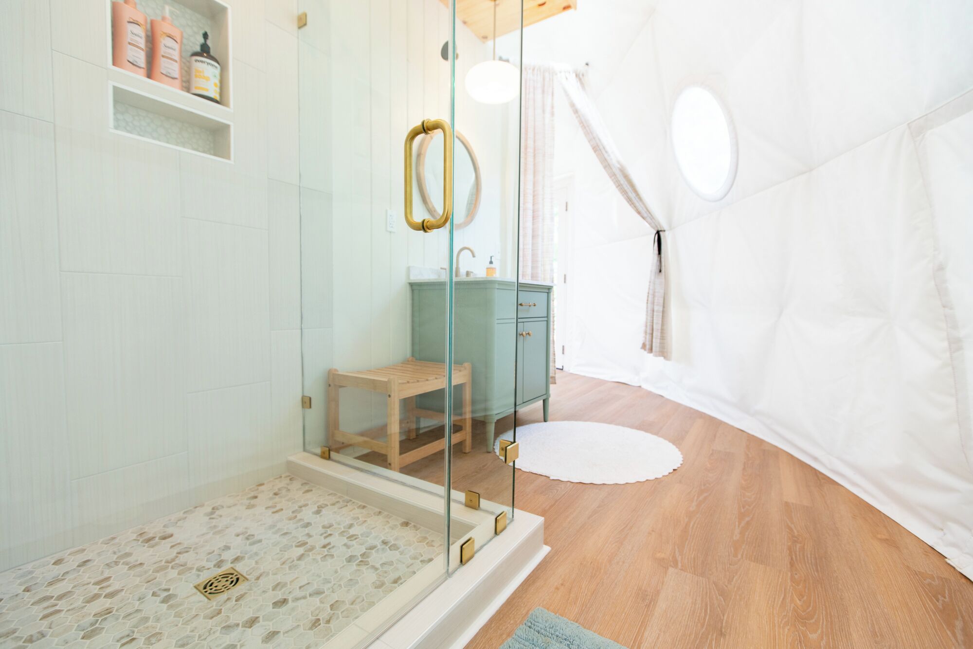 Un baño moderno con cabina de ducha de vidrio, un banco de madera, un mueble de tocador verde y una cortina blanca.