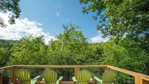 A wooden deck with four green chairs and a railing, overlooking a dense forest.