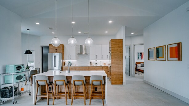 Una cocina moderna con una isla central, taburetes de bar, luces colgantes y un comedor con una mesa blanca.