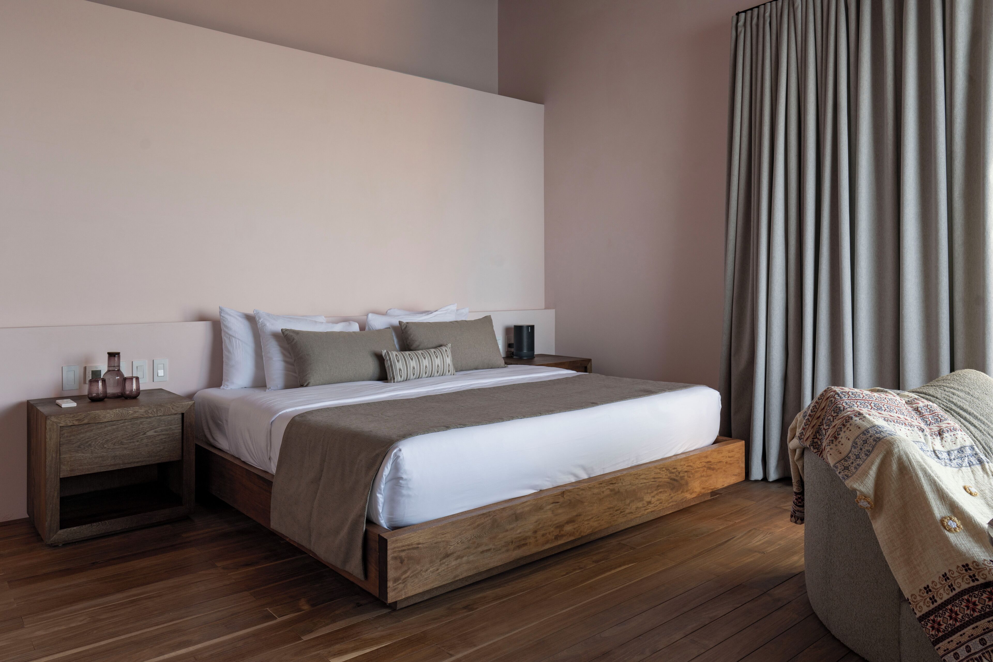 Un dormitorio moderno con estructura de cama de madera, una mesita de noche con un jarrón y un ventanal con cortinas.