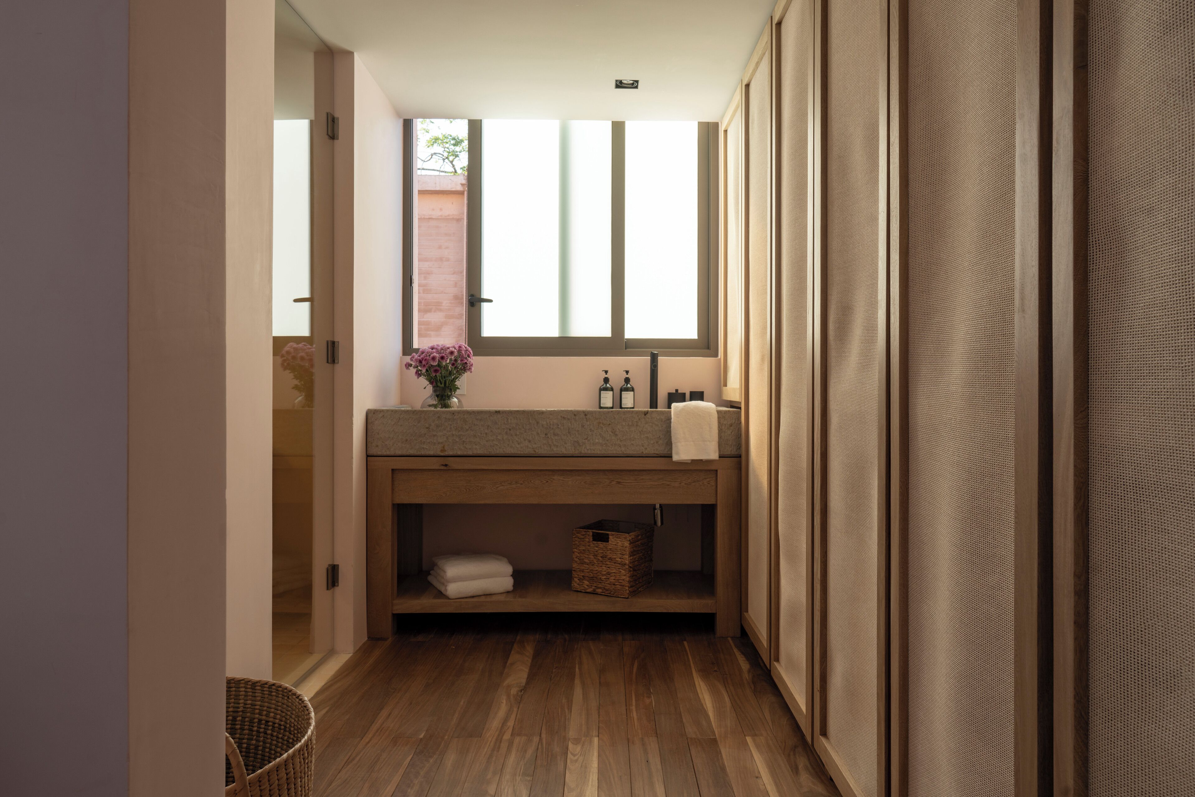 A narrow bathroom with a wooden vanity, a window, and a small shelf with towels.