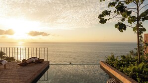 Une terrasse en bois avec une piscine à fond de verre donnant sur la mer, un banc en bois et un panier tressé.