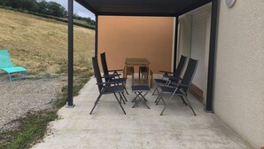 A covered patio with a table and chairs, a blue lounge chair, and a view of a hill. - New house with all comforts! (Ajac)