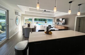 A modern kitchen with a central island, a dining area with a pool view, and a living room with a TV. - Cardinal - The Glass House (Canaan)