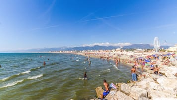 Una spiaggia affollata con persone che nuotano e prendono il sole, una ruota panoramica e montagne sullo sfondo.