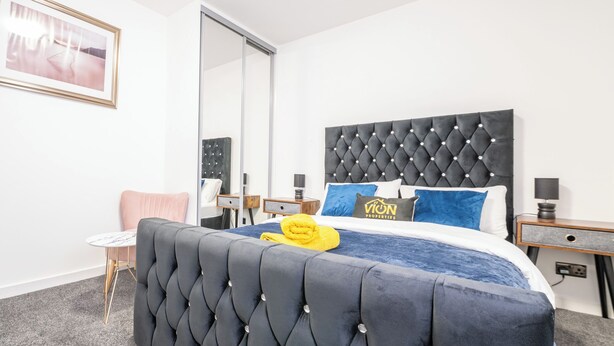 Une chambre à coucher avec une tête de lit capitonnée, un lit avec une literie bleue, une serviette jaune et un tableau encadré au mur.
