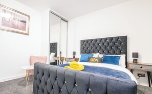 Un dormitorio con cabecera capitonada, cama con ropa de cama azul, una toalla amarilla y un cuadro enmarcado en la pared.